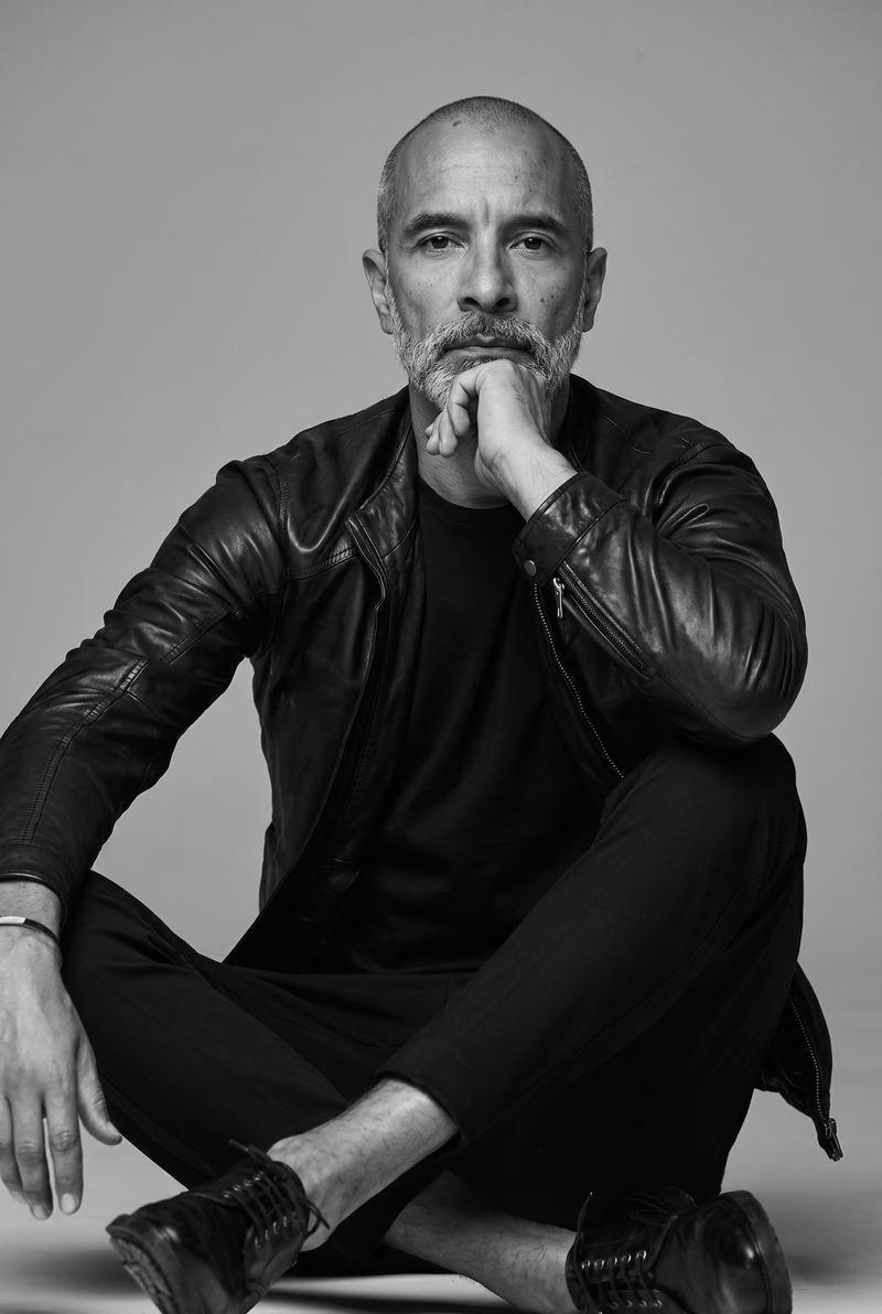 Pacho Sánchez sentado en el piso con chaqueta de cuero negra y la mano en la barbilla, retrato en blanco y negro